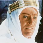 Ein Mann in weißer Wüstenkleidung mit intensiv blauen Augen schaut nach vorn. Er trägt einen goldenen Kopfschmuck.