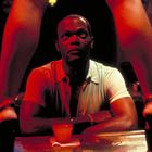 Ein Mann sitzt an einer Bar, zwischen ihm und der Kamera sind Beine in High Heels zu sehen. Er schaut konzentriert.