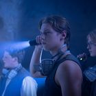 Drei Personen mit Atemmasken erkunden mit Taschenlampen einen dunklen Raum. Sie wirken angespannt und konzentriert.