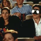 Eine Frau hält eine Popcorn-Tüte, daneben sitzt ein Mann mit seltsamer Brille in einem Kinosaal.