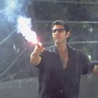 Ein Mann hält eine Leuchtrakete, trägt eine Brille und ein schwarzes Hemd. Hintergrund ist regnerisch und bewaldet.