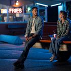 Leuchtschrift: "FRIES". Ein Mann und eine Frau sitzen auf einem Pickup-Truck vor einem Diner, trinken aus Pappbechern.