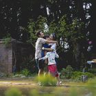 Drei Personen umarmen sich herzlich im Garten, umgeben von Bäumen und einem Ziegelbau im Hintergrund.