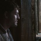 Ein Mann in einem dunklen Raum blickt nach draußen. Im Hintergrund sind zwei weitere Personen in einer Holzkulisse zu sehen.