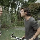 Zwei Männer stehen sich gegenüber, einer hält ein Fahrrad. Sie befinden sich in einem Garten mit Bäumen und Sträuchern.