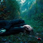 Ich kann den Text nicht erkennen. Eine Person liegt im Wald auf einem grasigen Pfad, umgeben von Bäumen und Herbstlaub.