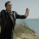 Ein Mann in Lederjacke mit erhobener Hand und Pistole, am Ufer eines Sees, wirkt besorgt und gestikuliert.