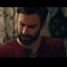 Ein Mann mit Bart in rotem Shirt schaut nachdenklich. Im Hintergrund ein Vorhang mit Blattmuster und unscharfe Dekoration.