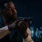 Ein Mann mit Bart zielt mit einer Pistole, trägt ein weißes Hemd und dunkle Weste. Hintergrund ist eine verschwommene Stadtansicht bei Nacht.