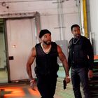 Zwei Männer in schwarzer Kleidung und Schutzwesten, einer hält eine Pistole. Umgeben von industrieller Umgebung.