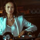 Eine Frau in einem eleganten weißen Blazer sitzt in einem gediegenen Raum, Blick direkt nach vorne, trägt eine Halskette.
