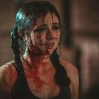 Eine Frau mit Zöpfen, bedeckt mit einer roten Substanz, wirkt verängstigt und emotional in einem düsteren Raum.