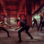 Es gibt keinen sichtbaren Text. Zwei junge Männer stehen in Kampfposition, umgeben von sechs anderen in einem städtischen Umfeld.