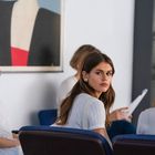 Ich kann die Texte nicht erkennen. Eine Gruppe von Menschen sitzt, eine Frau schaut aufmerksam, im Hintergrund modernes Gemälde.