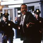 Ein Mann in Anzug hält die Arme aus, zwei Polizisten mit Waffen im Hintergrund, belebte, städtische Szenerie.