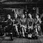 Fünf Frauen in Uniformen sitzen in einer Holzhütte. Sie tragen militärische Kleidung und Stiefel, einige halten Gewehre.