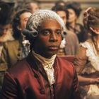 Ein Mann in historischer Kleidung mit gepuderter Perücke sitzt in einer Menschenmenge, die ebenfalls elegant gekleidet ist.