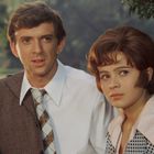 Eine Frau mit gepunkteter Bluse und ein Mann in Anzug stehen im Grünen, umgeben von Bäumen.