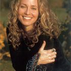 Keine sichtbaren Texte. Eine lächelnde Frau mit lockigem Haar in natürlicher Umgebung. Sie trägt einen Ring und eine verzierte Bluse.