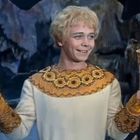 Ein lächelnder Mann hält einen bemalten Eimer in einer Hand, im Hintergrund eine mystische Höhle.