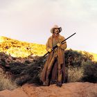 Ein Cowboy in langer Jacke und Hut steht in einer Wüstenlandschaft mit Felsen im Hintergrund.
