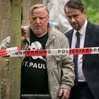 "POLIZEIABSPERRUNG" steht auf einem rot-weißen Absperrband. Zwei Männer, einer im Anzug, sind in einem Wald.