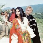 Ich kann keine Texte in dem Bild erkennen. Eine Frau neben einem Mann in historischer Kleidung, mit einem Wagen und Hügeln im Hintergrund.