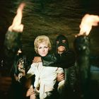 Keine sichtbaren Texte. Ein maskierter Mann hält eine Frau vor einer dunklen Höhlenwand, umgeben von Fackeln.