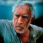Ein älterer Mann mit zerzaustem Haar und einem ernsten Gesichtsausdruck trägt ein zerschlissenes Hemd.