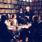 Ein Raum voller Bücherregale. Sechs sitzende Personen an einem Tisch mit Gläsern, eine stehende Person spricht.