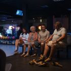 „BOWLING“, fünf Menschen in Bowlingschuhen sitzen in einem Bowlingcenter auf Bänken, bunte Kugeln im Hintergrund.