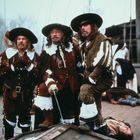 Drei Männer in historischer Kleidung, mit Hüten und Stiefeln, auf einem Schiff. Einer hält ein Schwert. Im Hintergrund Menschen.