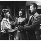 Eine junge Frau und ein Mann stehen sich gegenüber und halten Hände. Eine weitere Frau steht hinter ihnen und schaut zu.