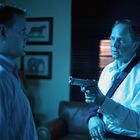 Links ein Mann in einem gestreiften Hemd, rechts ein Mann in einem weißen Hemd mit Pistole. Im Hintergrund Tierbilder.