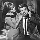 Ein Mann telefoniert lächelnd, während eine Frau ihn zärtlich umarmt. Beide wirken glücklich und elegant gekleidet.