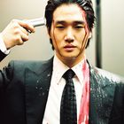 Es gibt keinen Text im Bild. Ein Mann in einem Anzug hält eine Pistole an seinen Kopf; Blut ist auf Gesicht und Anzug verteilt.