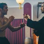 Ein junger, muskulöser Mann in Kampfhaltung gegenüber einem älteren, asiatischen Mann in schwarzer Kleidung, in einem Dojo.