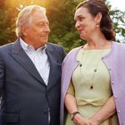 Ein älteres Paar steht lächelnd im Freien, in Sonnenlicht gehüllt, umgeben von Bäumen.