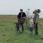 Keine erkennbaren Texte. Drei Personen: ein Mann mit Fahrrad, ein älterer Mann und ein Kind; am Ufer einer Wiese.