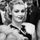Es gibt keinen Text. Eine Frau mit elegantem Styling, Blumen im Haar, trägt eine gemusterte Bluse und einen Pelz, lächelnd.
