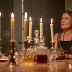 Eine elegant gekleidete Frau sitzt an einem Tisch mit Kerzenleuchtern, umgeben von Kristallgläsern und einer gemütlichen Atmosphäre.
