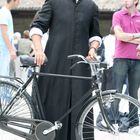 Mann in schwarzer Robe mit schwarzem Barett hält ein Fahrrad. Hintergrund zeigt mehrere Personen im Freien.