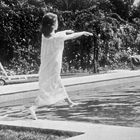 Eine Frau in einem langen weißen Kleid geht am Rand eines Pools entlang, hinter ihr ist dichte Vegetation und eine Liege.