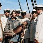 Männer in weißen Uniformen stehen auf einem Schiff. Einer hält einen erschöpften, dunkelhäutigen Jungen in den Armen.