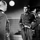 Drei Soldaten in Uniform in einem Raum; einer steht stramm, zwei andere sind locker. Hintergrund zeigt Regale mit Modellflugzeugen.