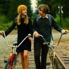 Zwei Jugendliche schieben Fahrräder auf einem alten Schienenweg, umgeben von grüner Natur, sie schauen sich an und lächeln.