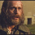 Ein Mann mit langem Haar und Bart schaut überrascht. Im Hintergrund ein unscharfes Gebäude mit Fenstern.