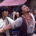 Eine Frau hält den Arm eines Mannes, als ob sie ihn stoppt. Beide tragen lässige, traditionelle Kleidung. Im Hintergrund sind weitere Personen und eine Hütte mit einem roten Dach zu sehen.