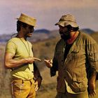 Zwei Männer in Freizeitkleidung stehen in einer hügeligen Landschaft und deuten mit den Fingern aufeinander.