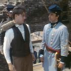 Zwei Männer in historischen Uniformen stehen; einer trägt blau mit knöpfen, der andere hat ein Seil um den Hals.